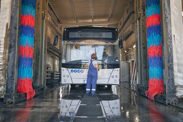 Bus passing through an automated vehicle wash system with rotating brushes, while a worker manually cleans the windscreen.