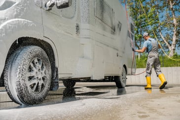 Operator cleaning a large commercial vehicle with foam and water at a commercial vehicle wash facility.